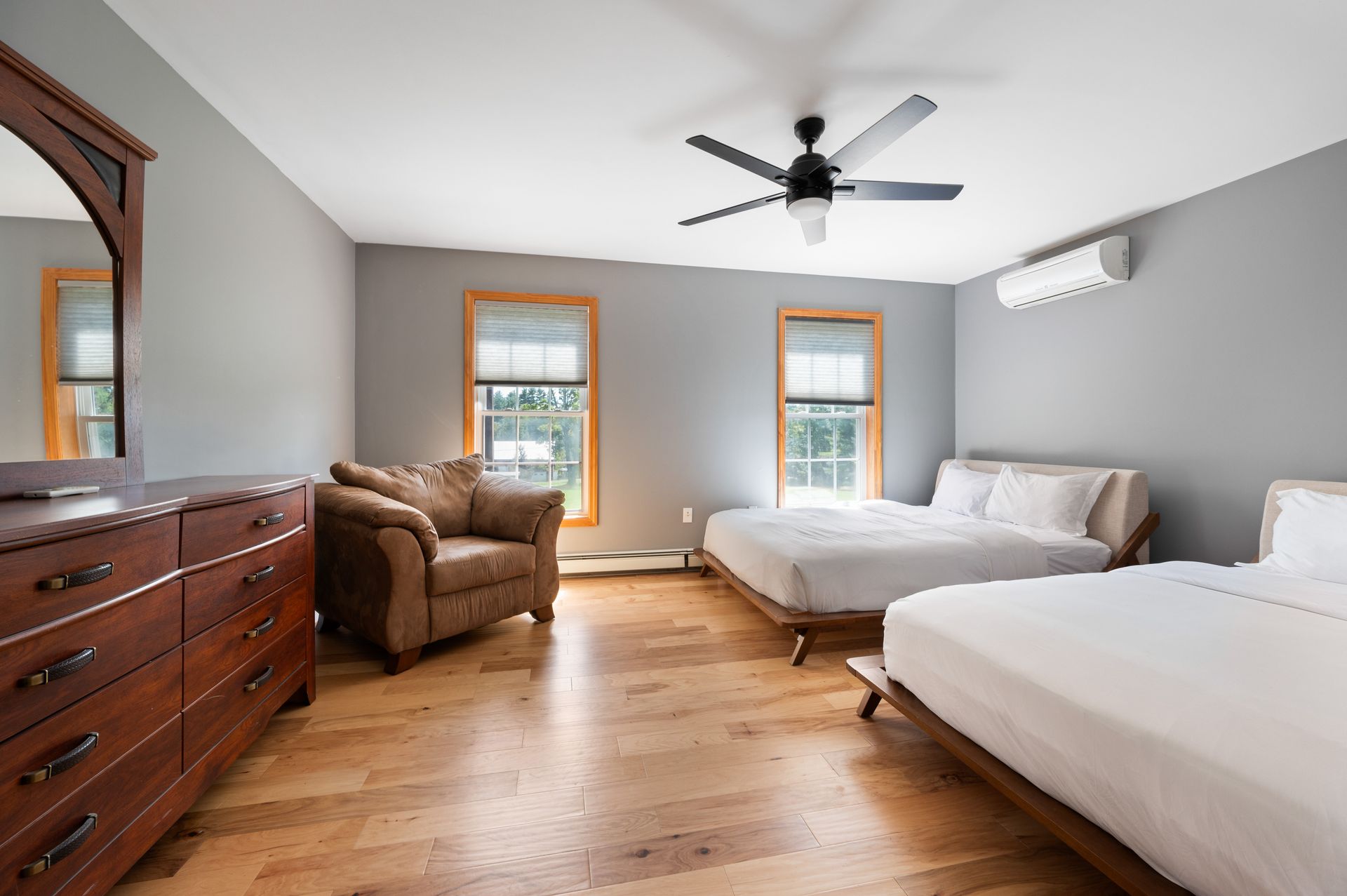 Bedroom with two beds, an armchair, and a dresser. Light-colored hardwood floor, gray walls, and natural light.