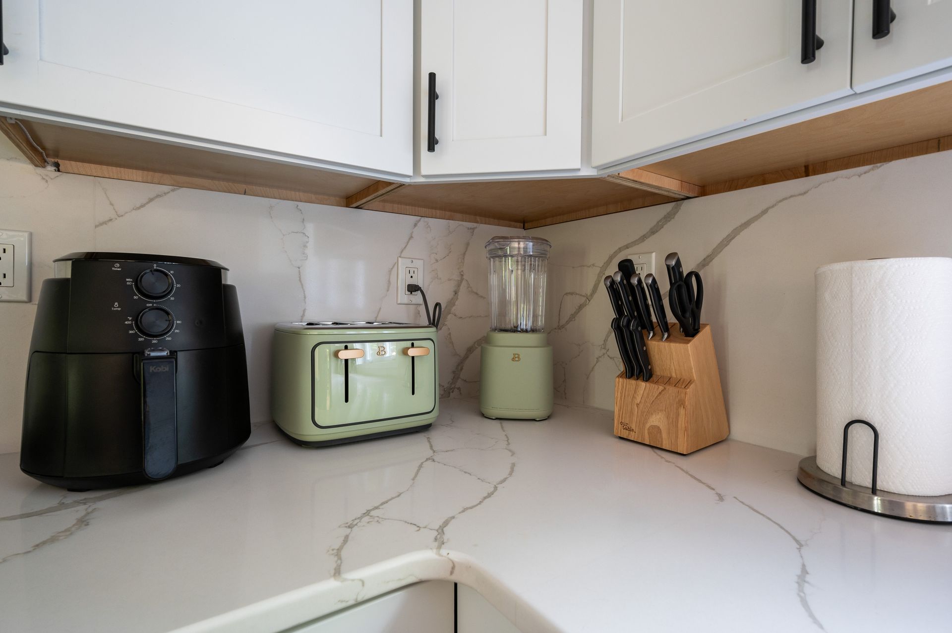 Kitchen countertop with appliances: air fryer, toaster, blender, knife block, and paper towel holder under white cabinets.