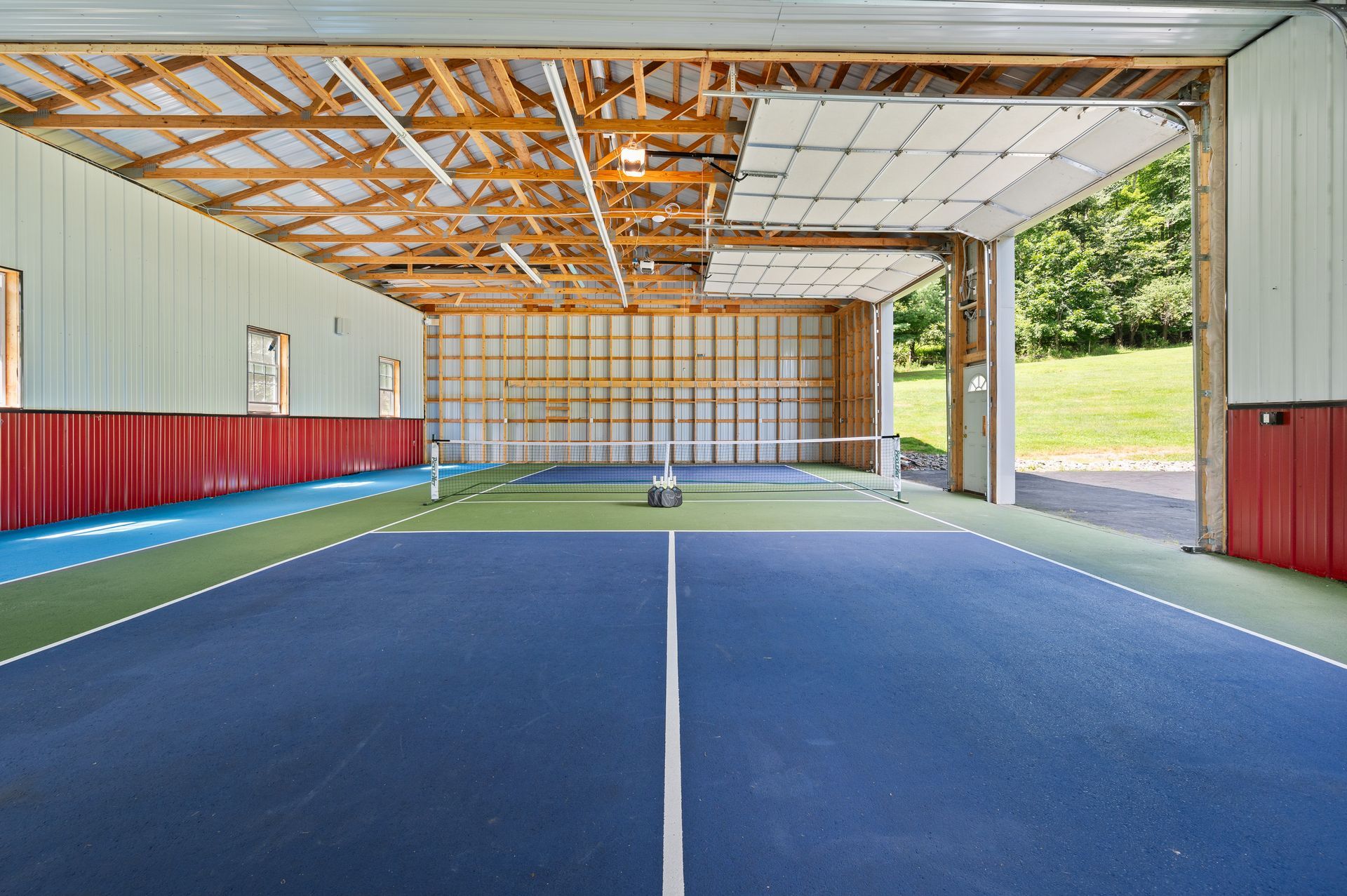 Indoor pickleball court with a blue and green playing surface, net, and open garage door.