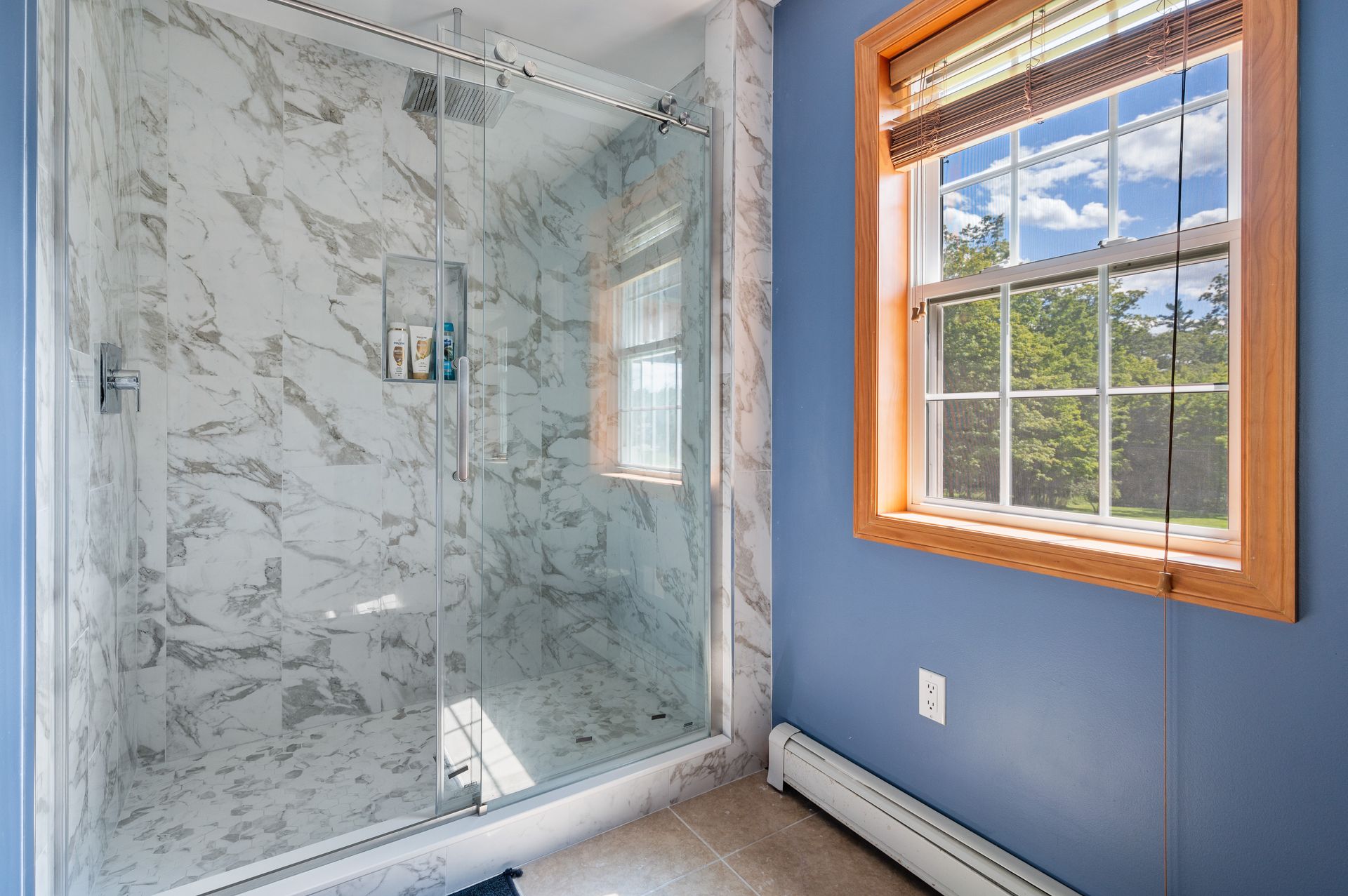 Modern bathroom with marble-tiled shower, glass doors, and a window with a view. Blue walls and wood trim.