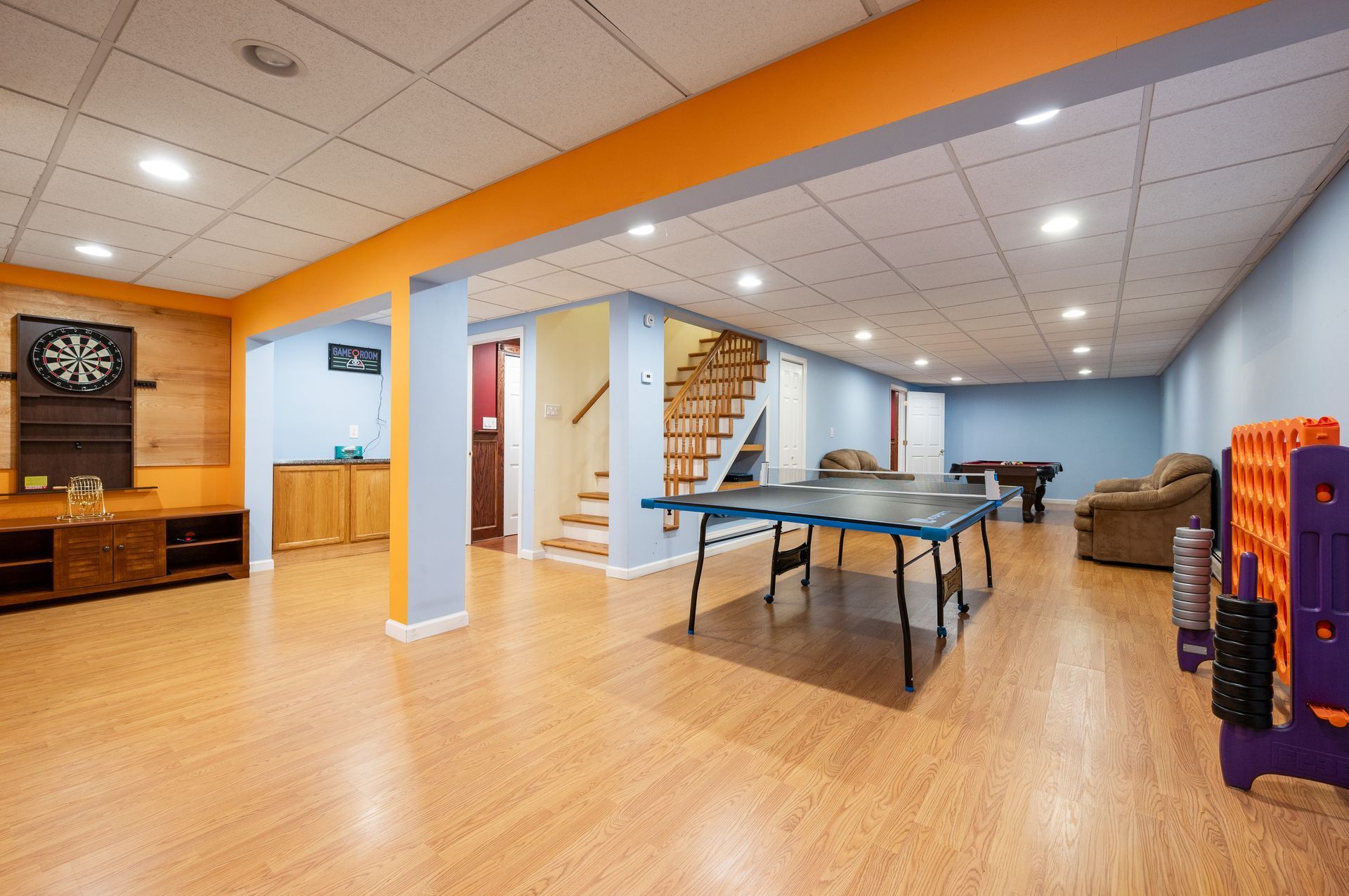 Spacious finished basement with ping pong table, dartboard, and stairs. Orange and blue walls, wood floor.