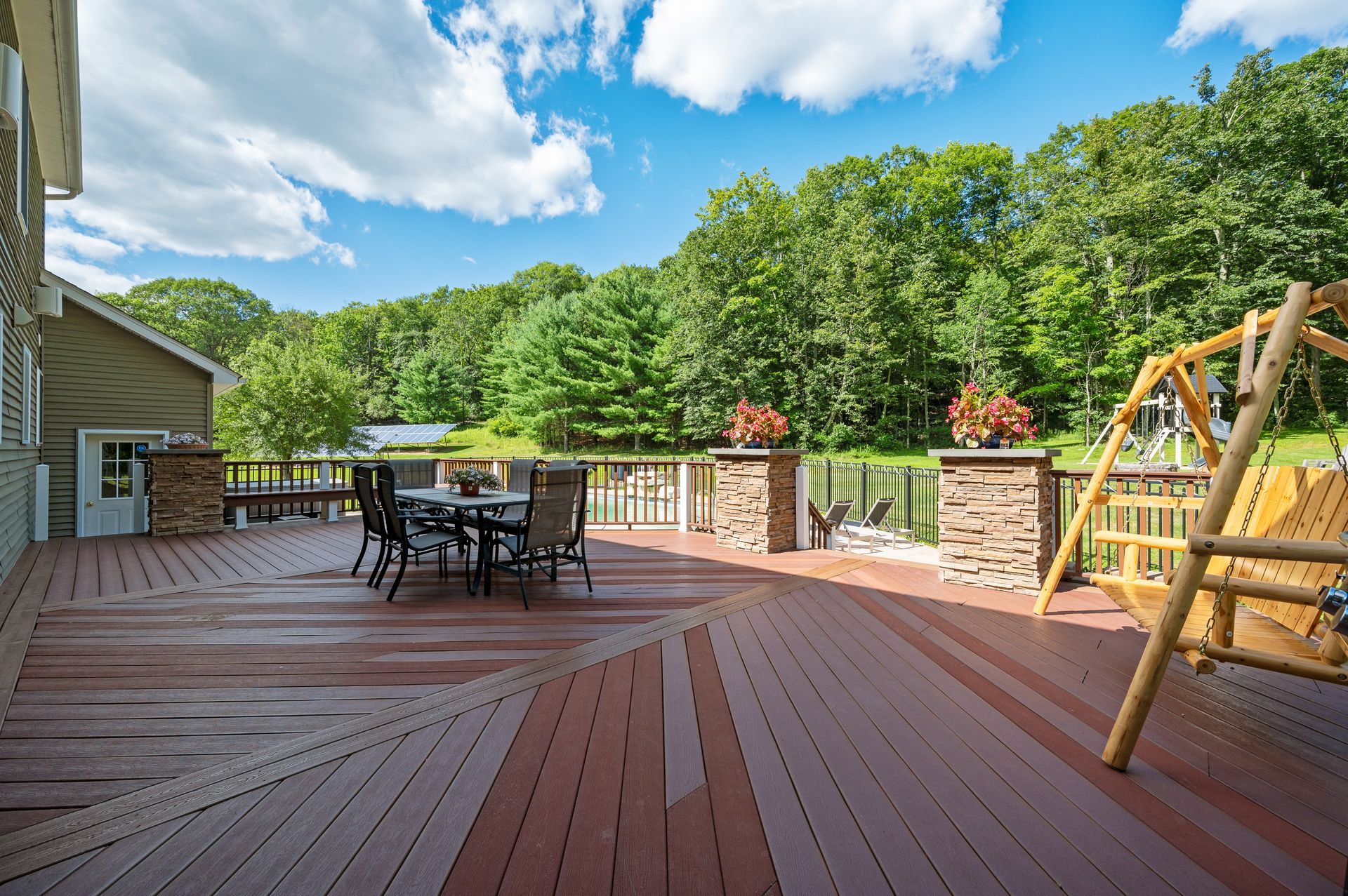 Wooden deck with dining table and swing overlooking a wooded area under a blue sky.