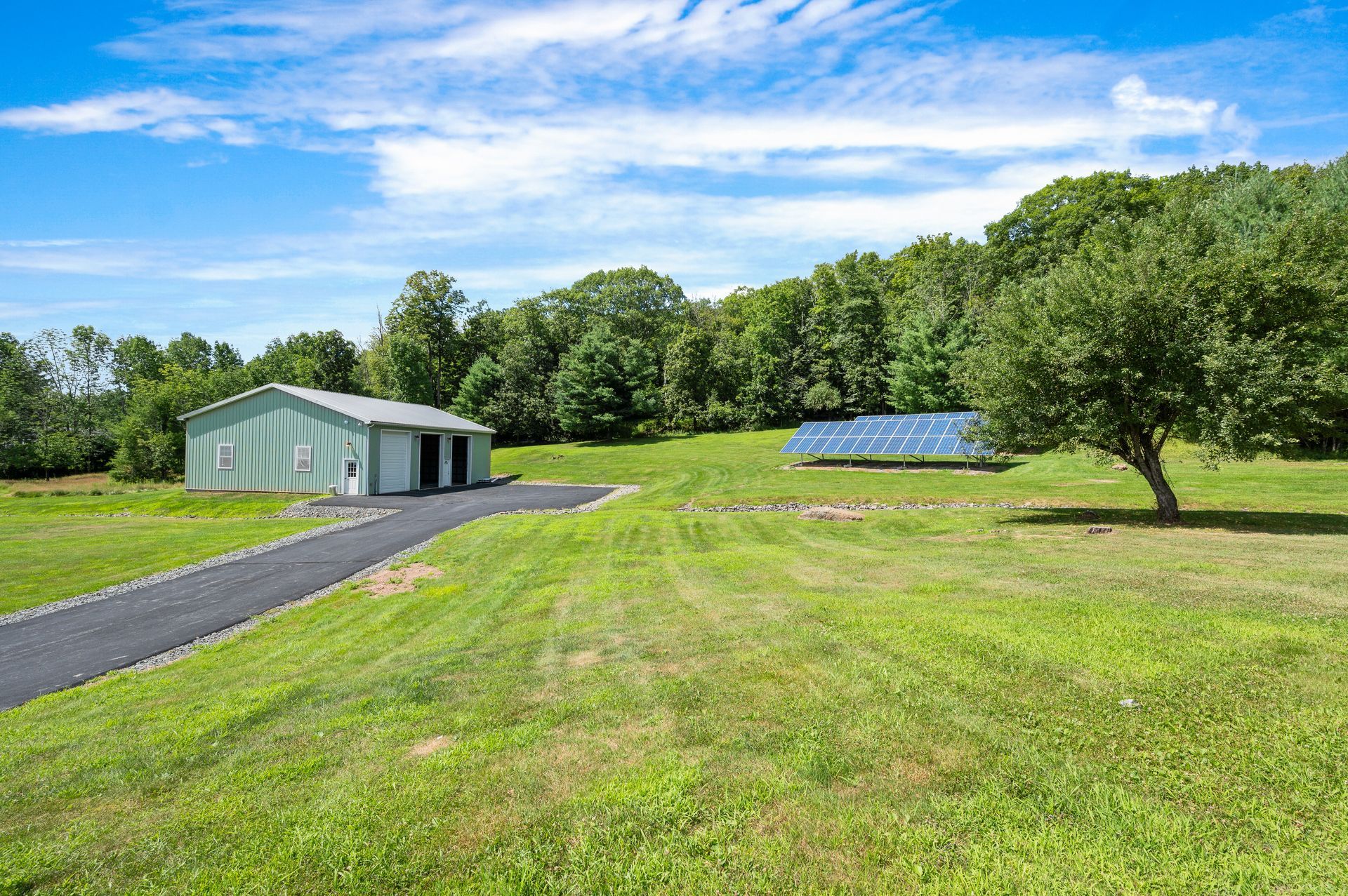 Green field with a building and a long, dark driveway leading to it; blue sky with trees in the background.