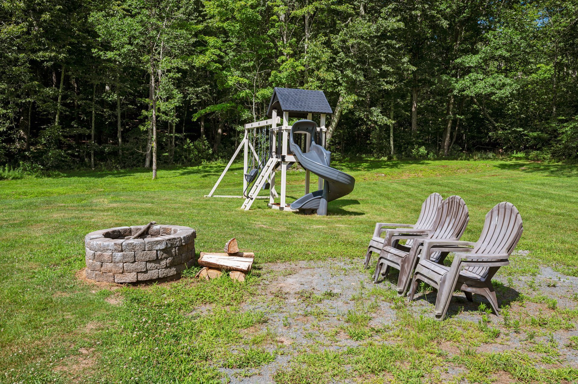 Backyard scene with a playset, fire pit, and Adirondack chairs on green grass.
