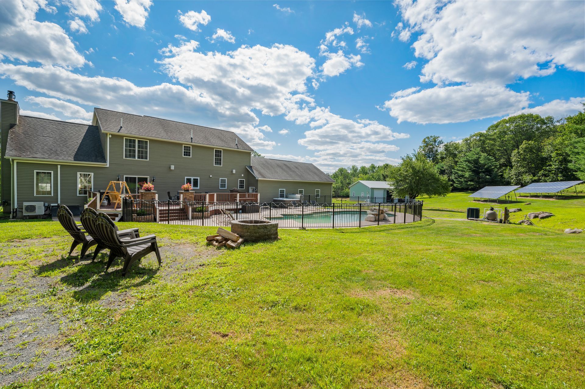 Green house with pool and yard under a blue sky with fluffy clouds. Two chairs sit on the grass.