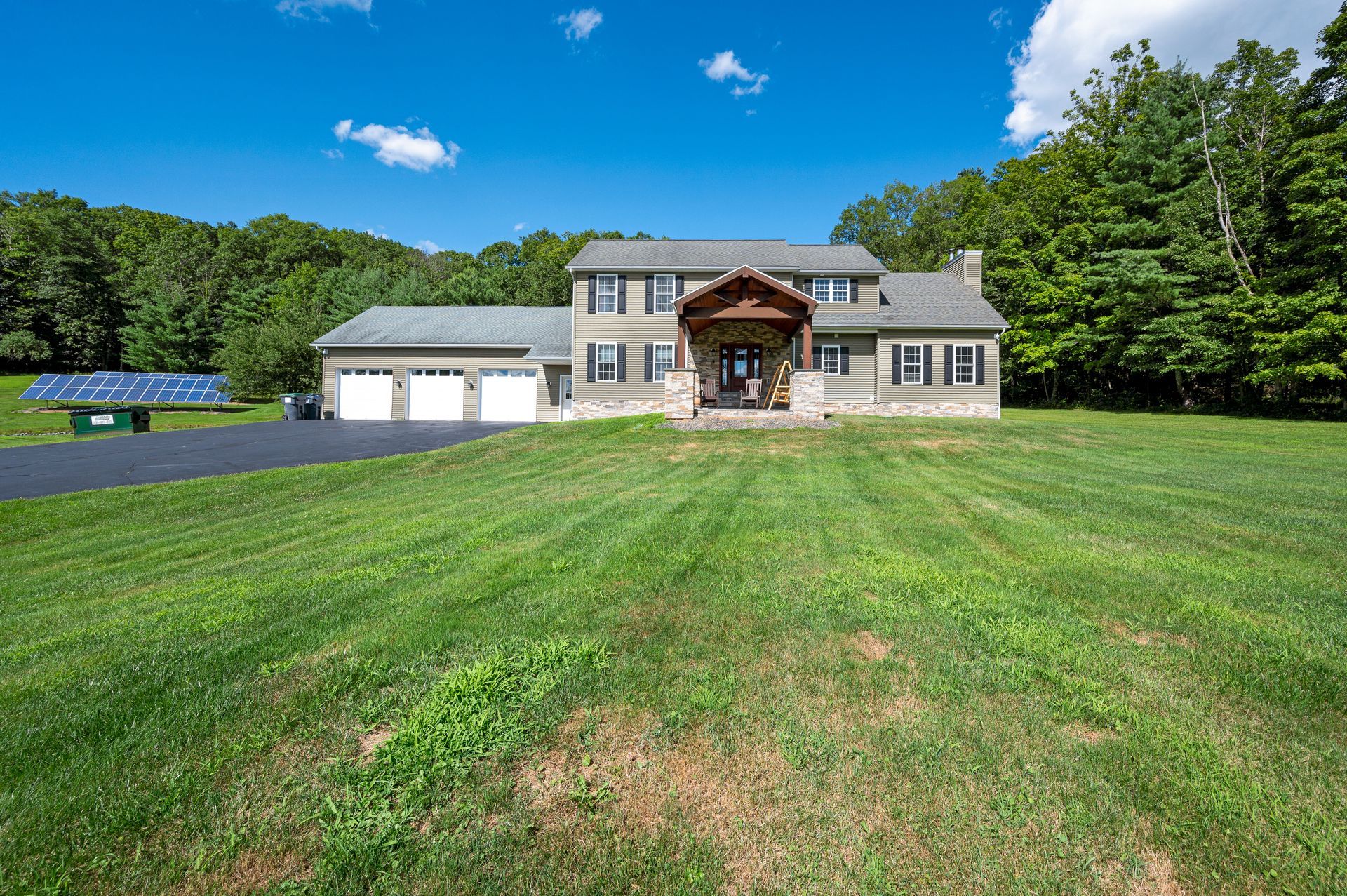 House with stone facade, two-car garage, and large lawn under a blue sky, surrounded by trees.