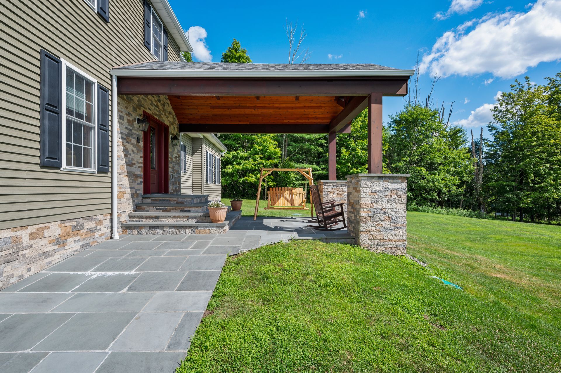Stone porch with dark wood beams, swing, and green lawn. House with gray siding and stone accents.