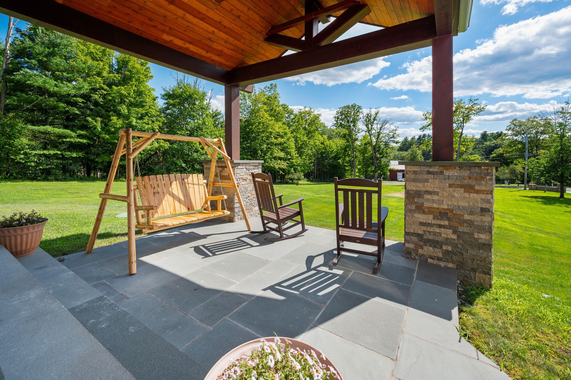 Covered porch with swing, rocking chairs, and stone-tiled floor, overlooking a green lawn under a sunny sky.