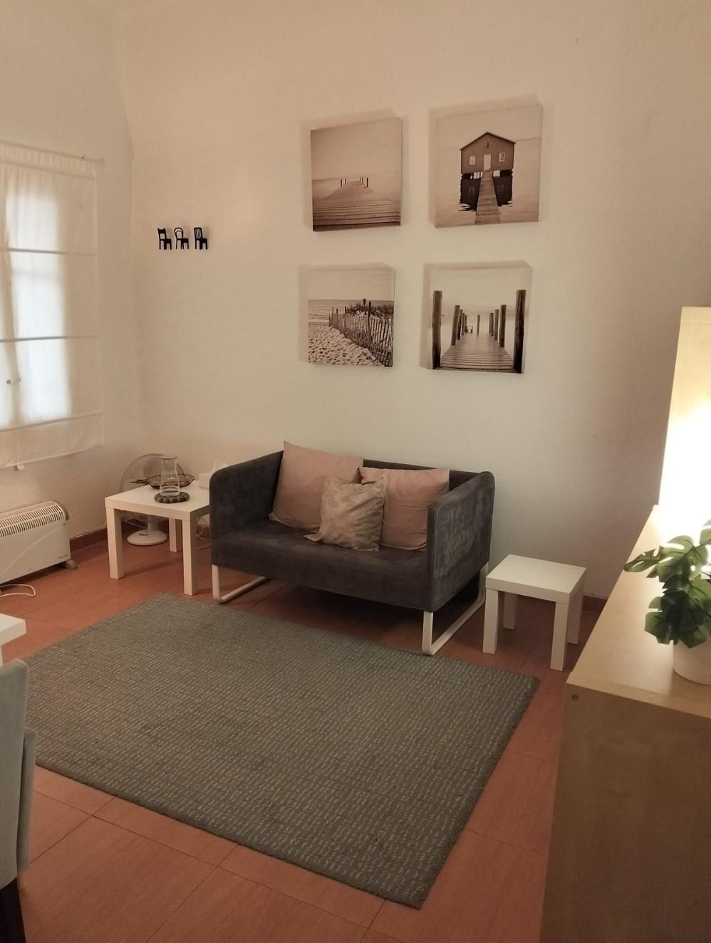 Una sala de estar con un sofá gris con cojines, dos mesitas auxiliares blancas, una alfombra gris y cuadros en la pared.