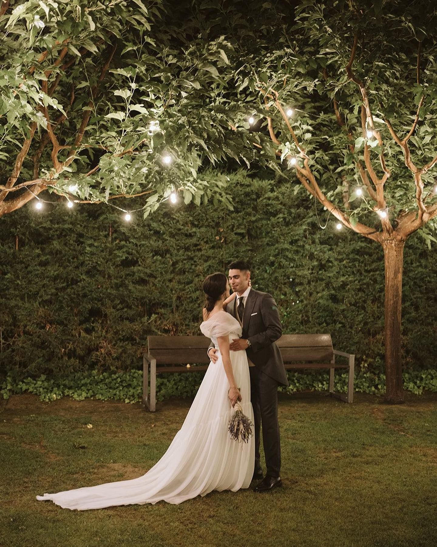 Una novia y un novio se besan bajo un árbol con luces colgando de él.