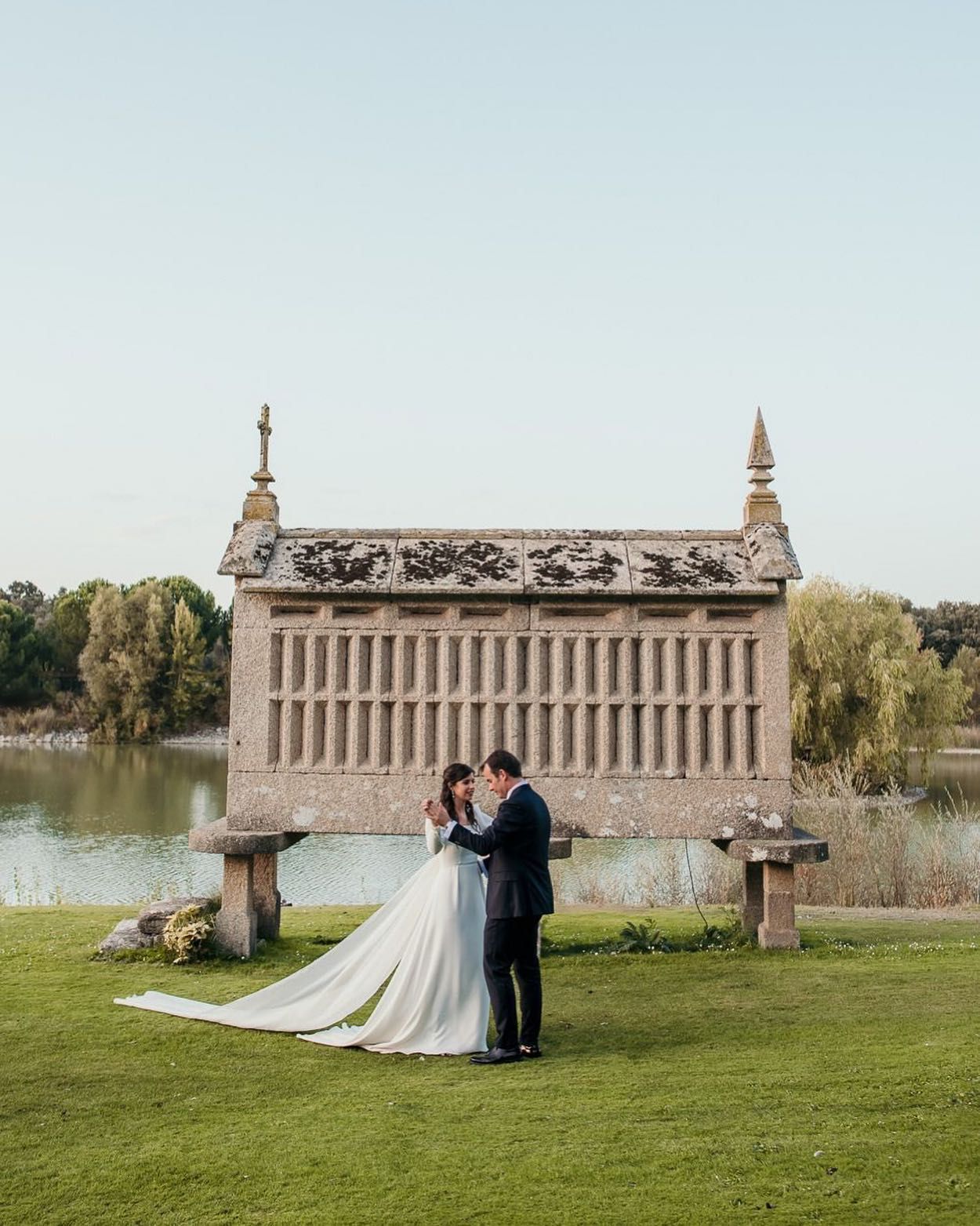 Una novia y un novio están de pie frente a un edificio al lado de un lago.