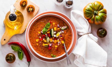 Un tazón de sopa está rodeado de verduras y especias sobre una mesa.