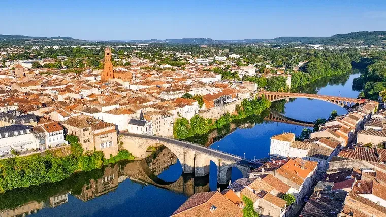 Vue aérienne d’une ville fluviale avec un pont en pierre, des toits orange et un clocher sous un ciel dégagé