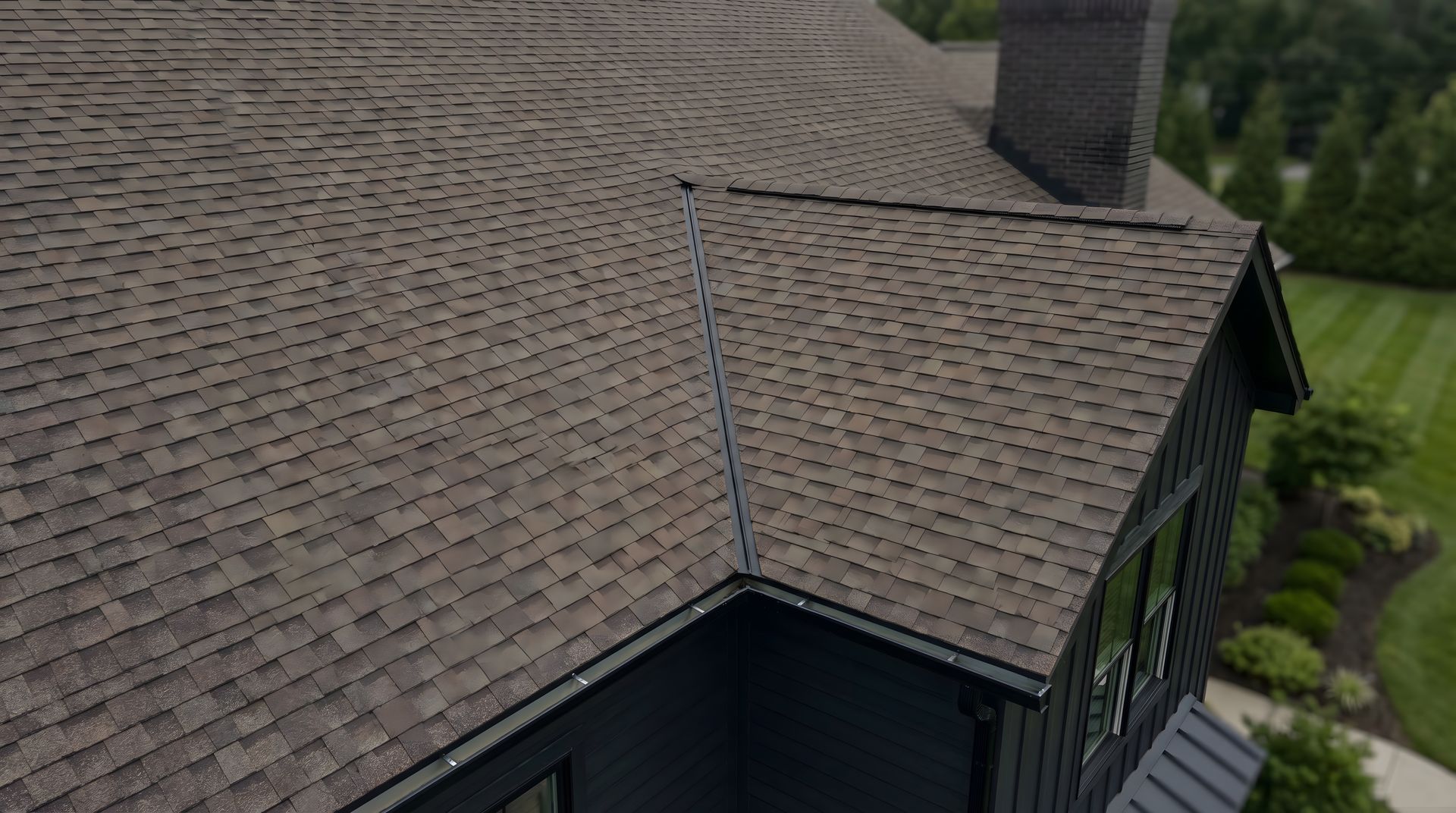 Vue aérienne d’un toit de maison brun recouvert de bardeaux, avec une cheminée et de la verdure autour.