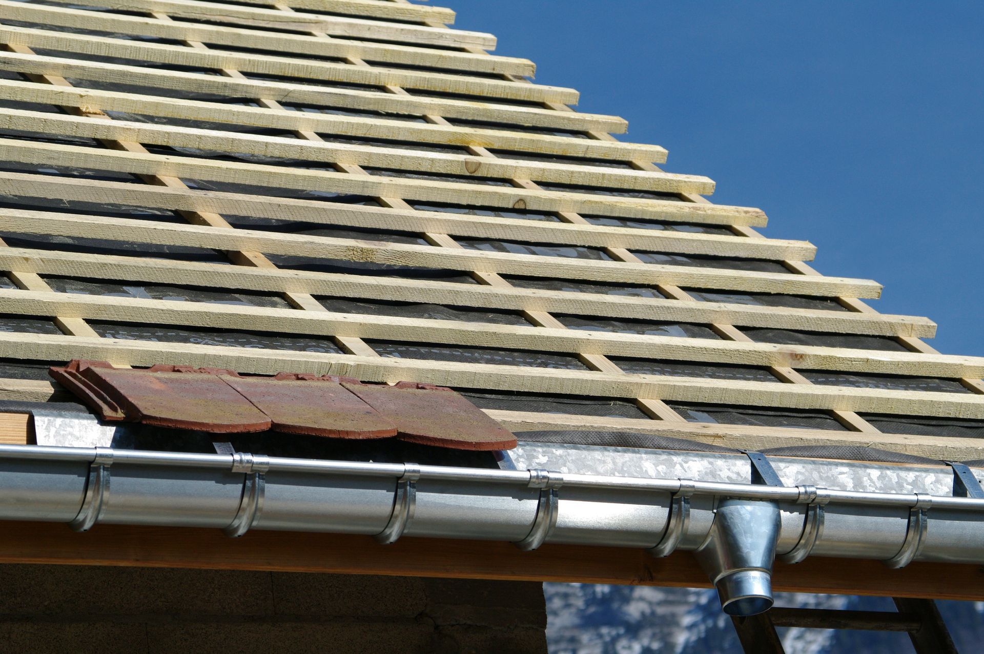 Toit en construction avec des chevrons en bois, une gouttière argentée et un ciel bleu en arrière-plan