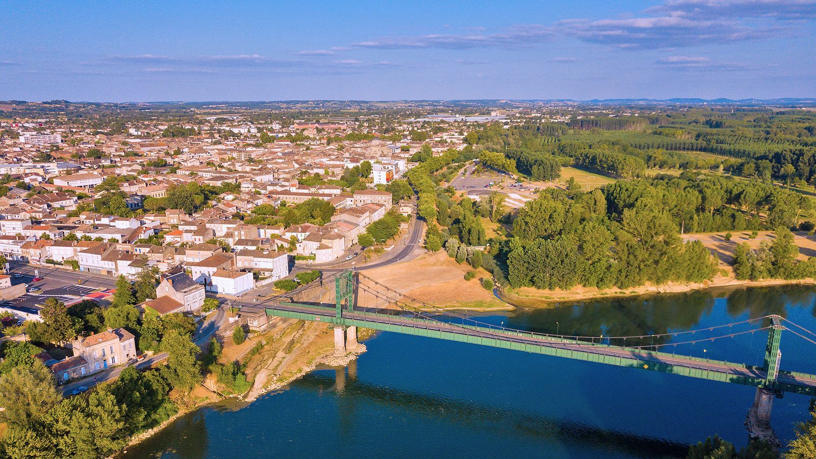 Vue aérienne d’une ville au bord d’une rivière, avec un pont, une eau bleue et des collines verdoyantes sous un ciel dégagé