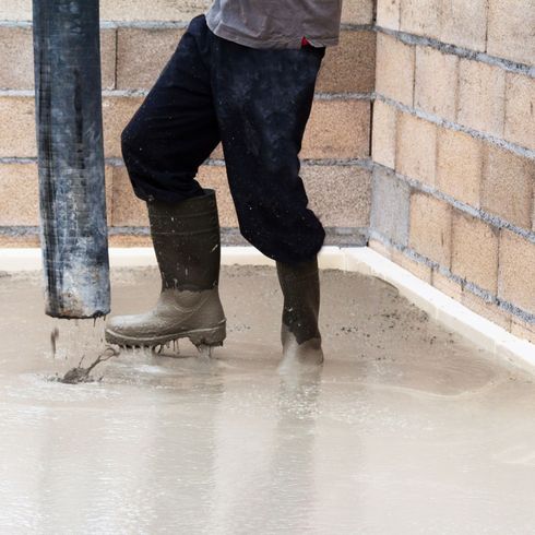Un ouvrier en bottes de caoutchouc coule du béton à côté d'un mur de briques et d'un poteau métallique.