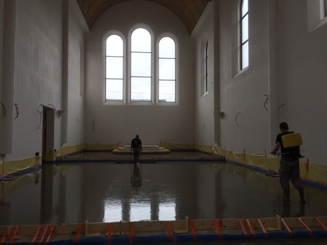 Salle de sport intérieure avec un sol brillant, de grandes fenêtres cintrées et deux personnes debout de part et d'autre.