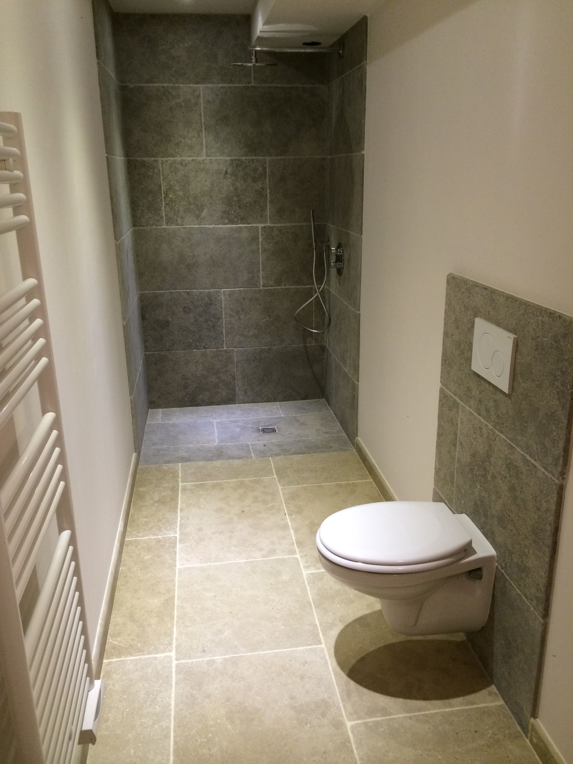 Salle de bains étroite carrelée avec toilettes suspendues et douche à l'italienne au fond.