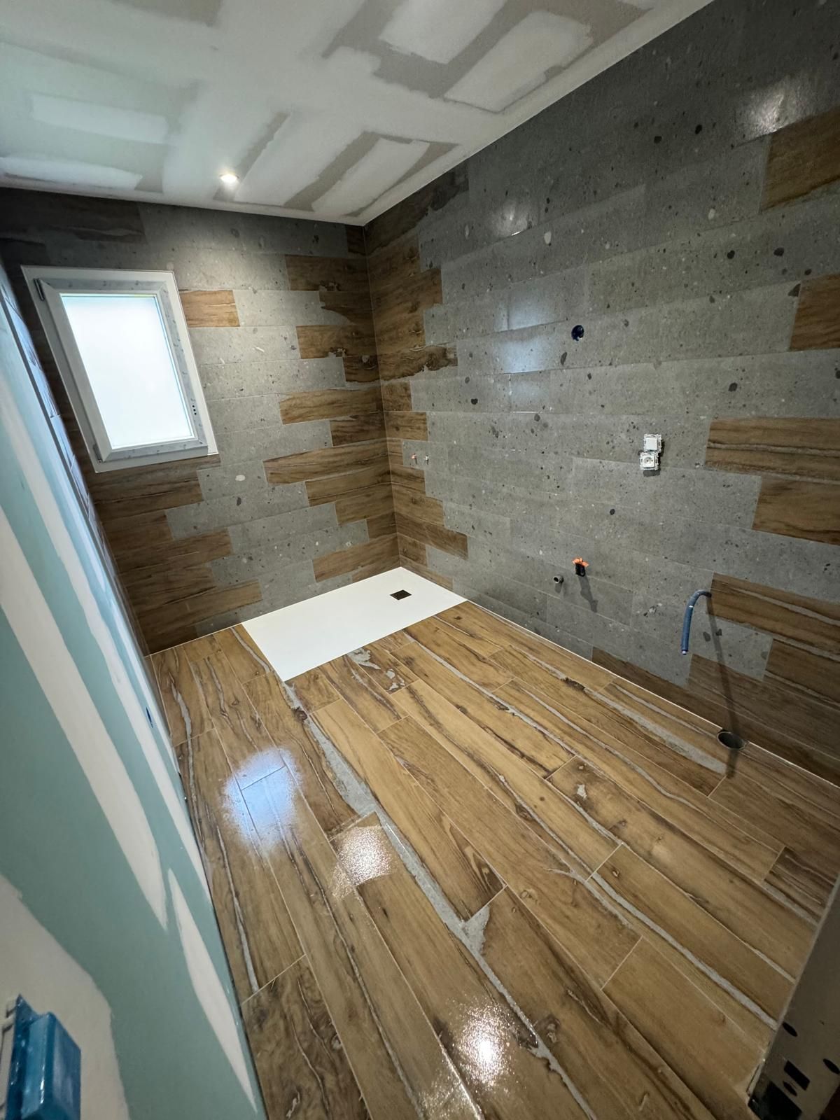 Petite salle de bains en cours de rénovation avec murs carrelés, sol imitation bois, fenêtre et receveur de douche.