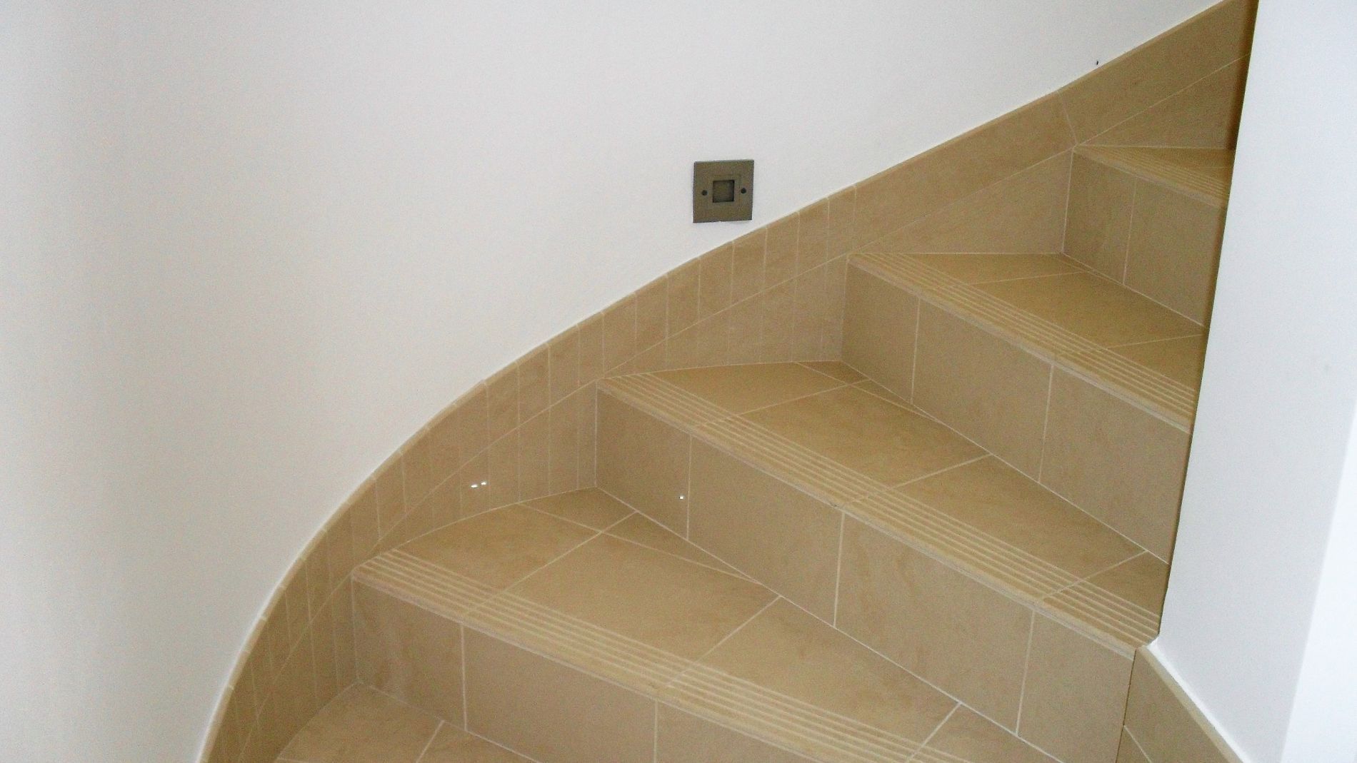 Escalier incurvé carrelé avec murs et une petite prise murale à proximité.