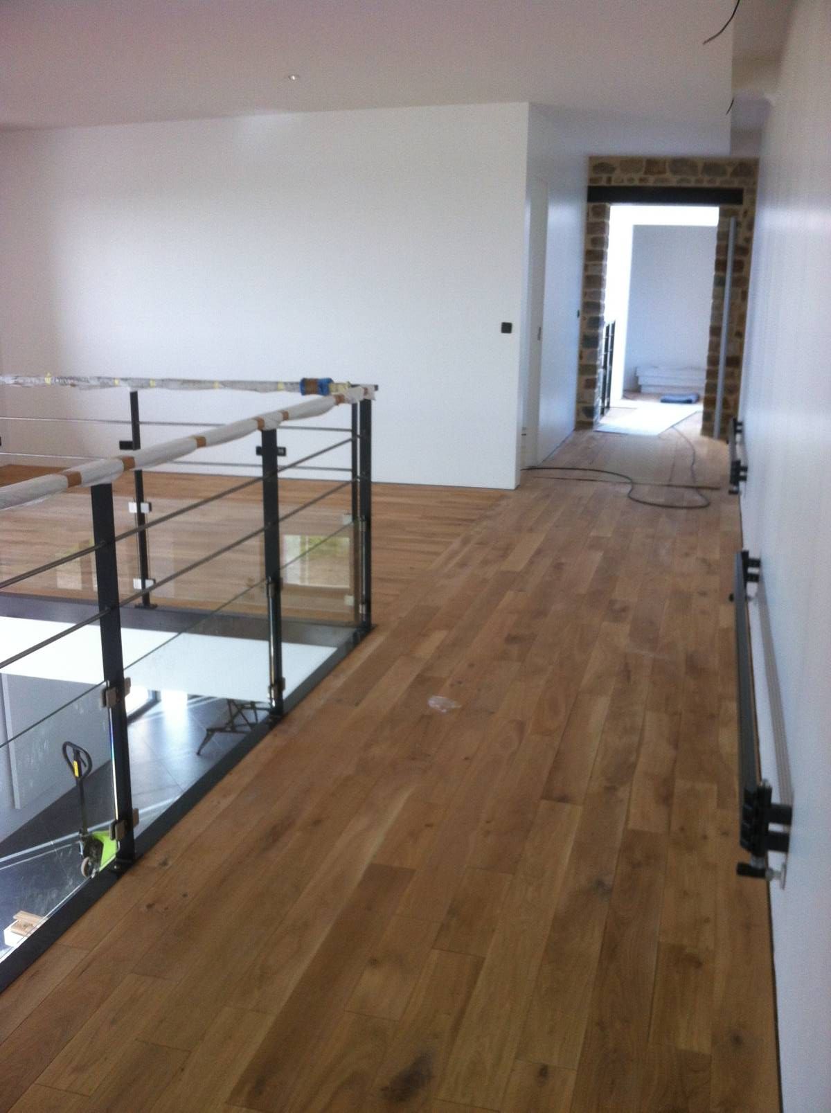 Couloir moderne vide avec parquet, rambarde en verre et porte au fond.