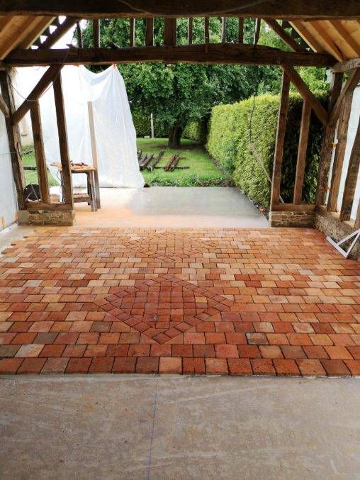 Terrasse en briques sous une pergola en bois, avec un rideau et un jardin bordé d'une haie au-delà.