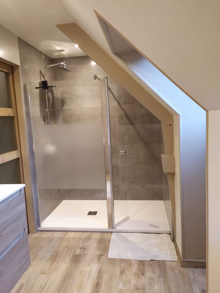 Salle de bains moderne avec douche en verre, carrelage et plafond mansardé sous un puits de lumière.