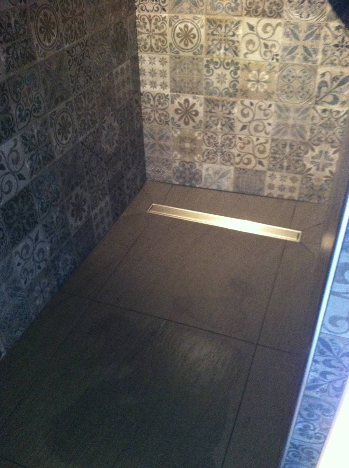 Petite cabine de douche carrelée avec un sol sombre et une bande de soleil qui la traverse.