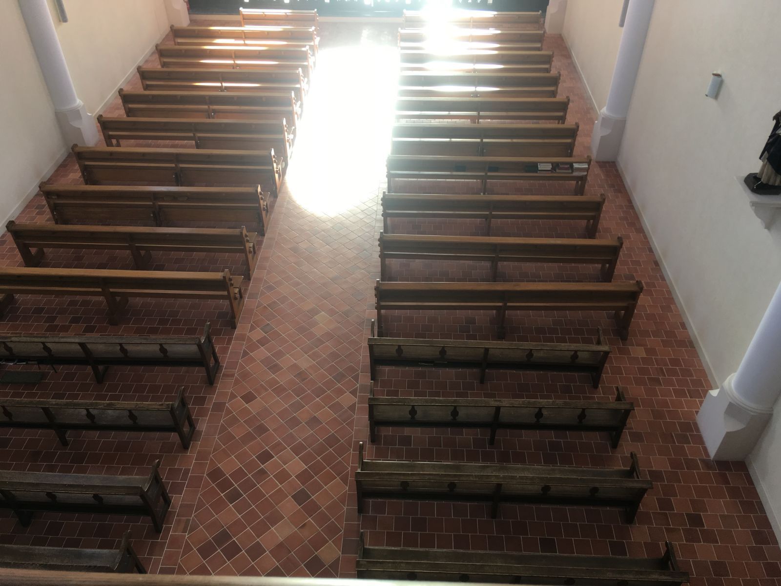 Des bancs d'église vides font face à une porte lumineuse dans une allée de chapelle au sol de briques.