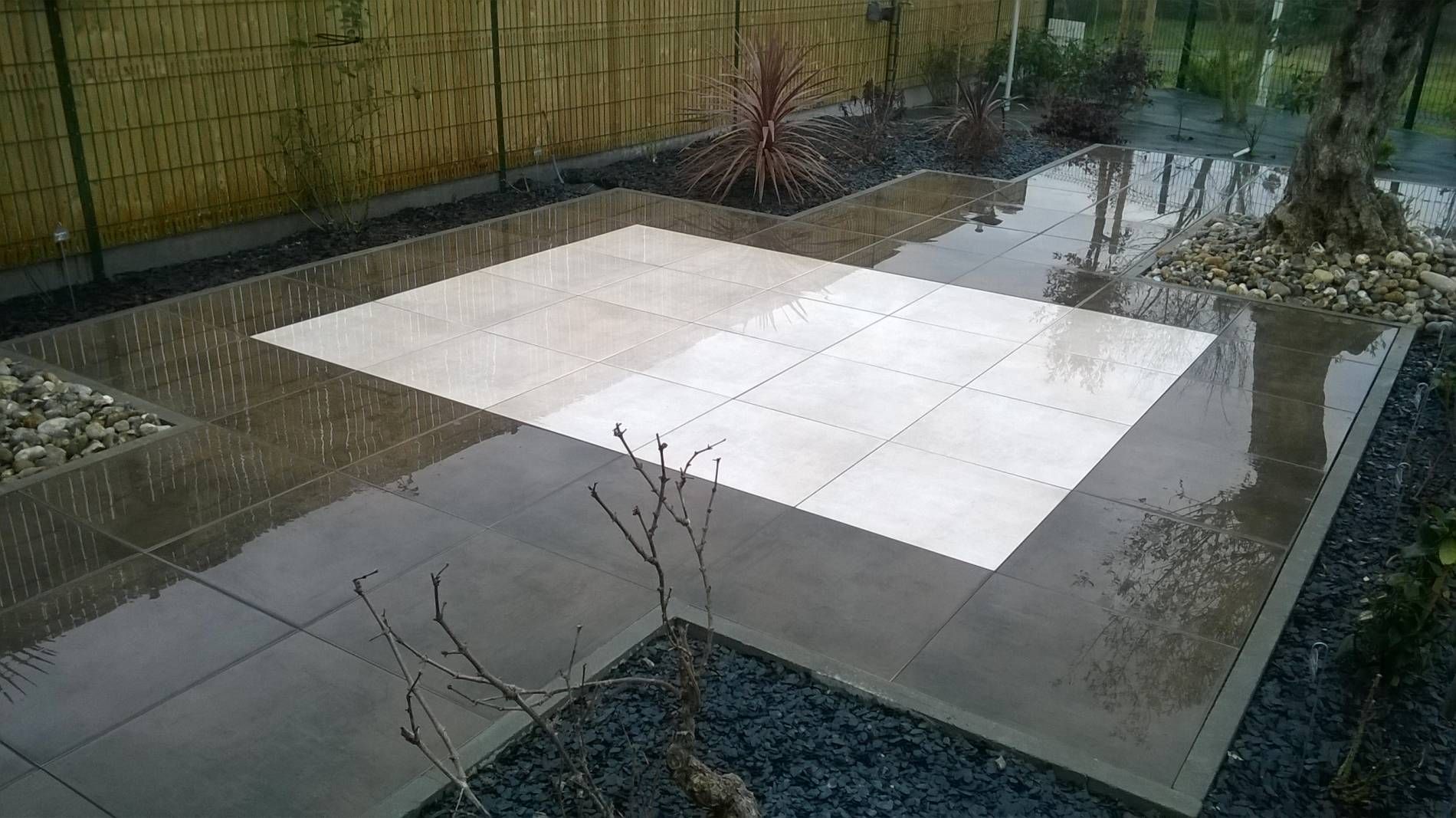 Dalles de terrasse carrées, vues du dessus, le long d'un chemin de jardin humide et d'une clôture.