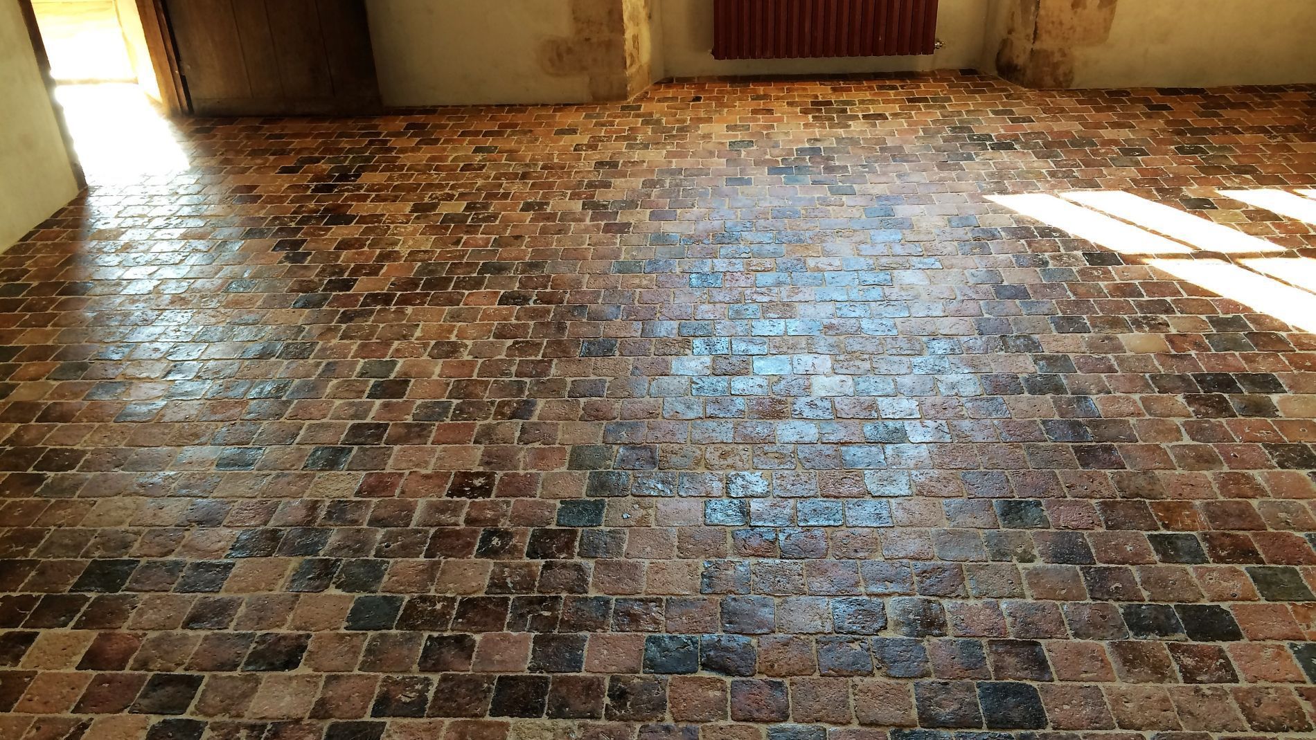 Sol en briques baigné de soleil dans une pièce intérieure vide, avec une porte et un radiateur le long du mur.