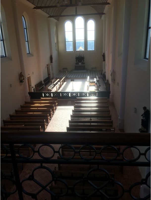 Intérieur d'église baigné de soleil, avec des bancs en bois faisant face à un autel et de hautes fenêtres cintrées.