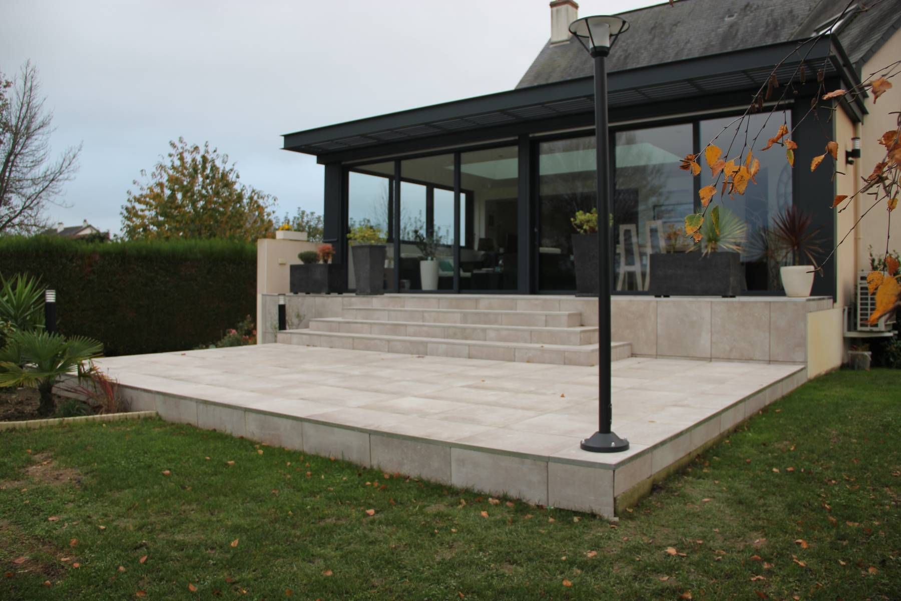 Patio avec marches en pierre surélevées, poteaux de pergola et façade de maison avec portes vitrées.