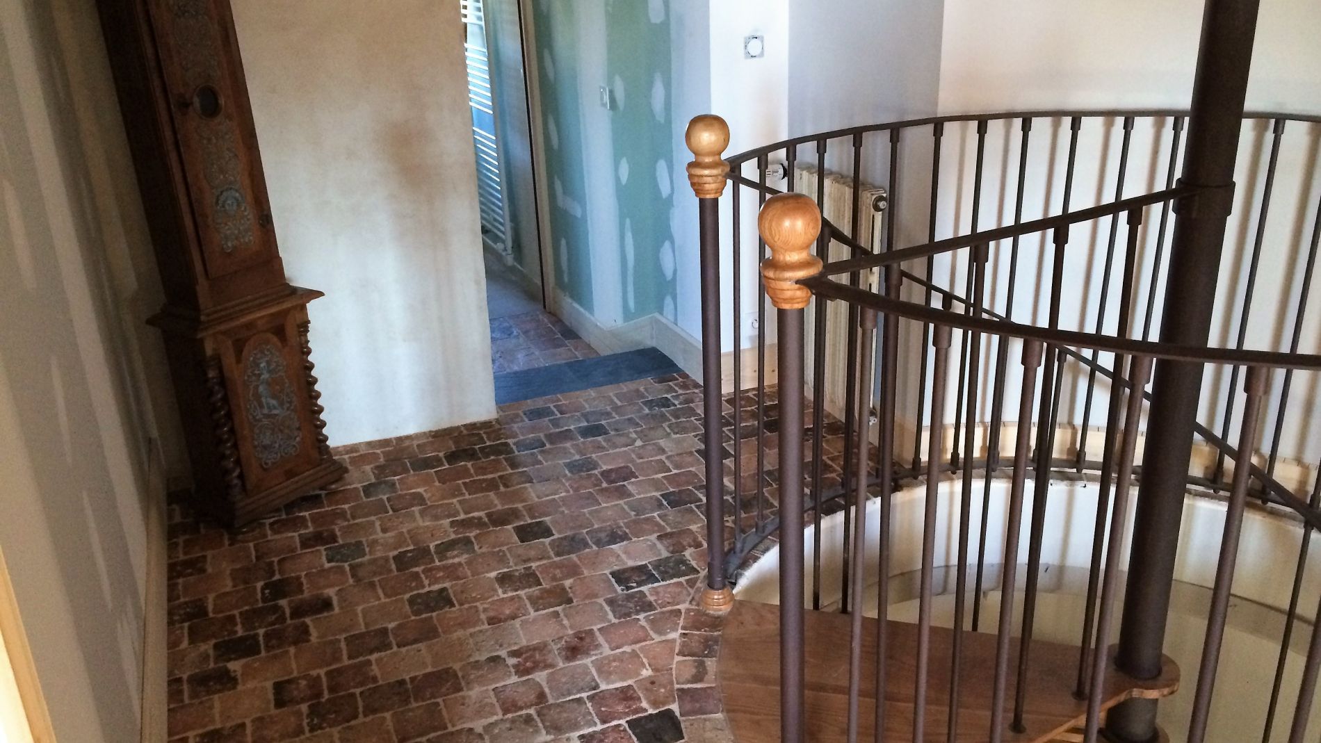Couloir intérieur avec sol carrelé à motif de briques, rampe d'escalier courbe et porte ouverte donnant sur un hall.