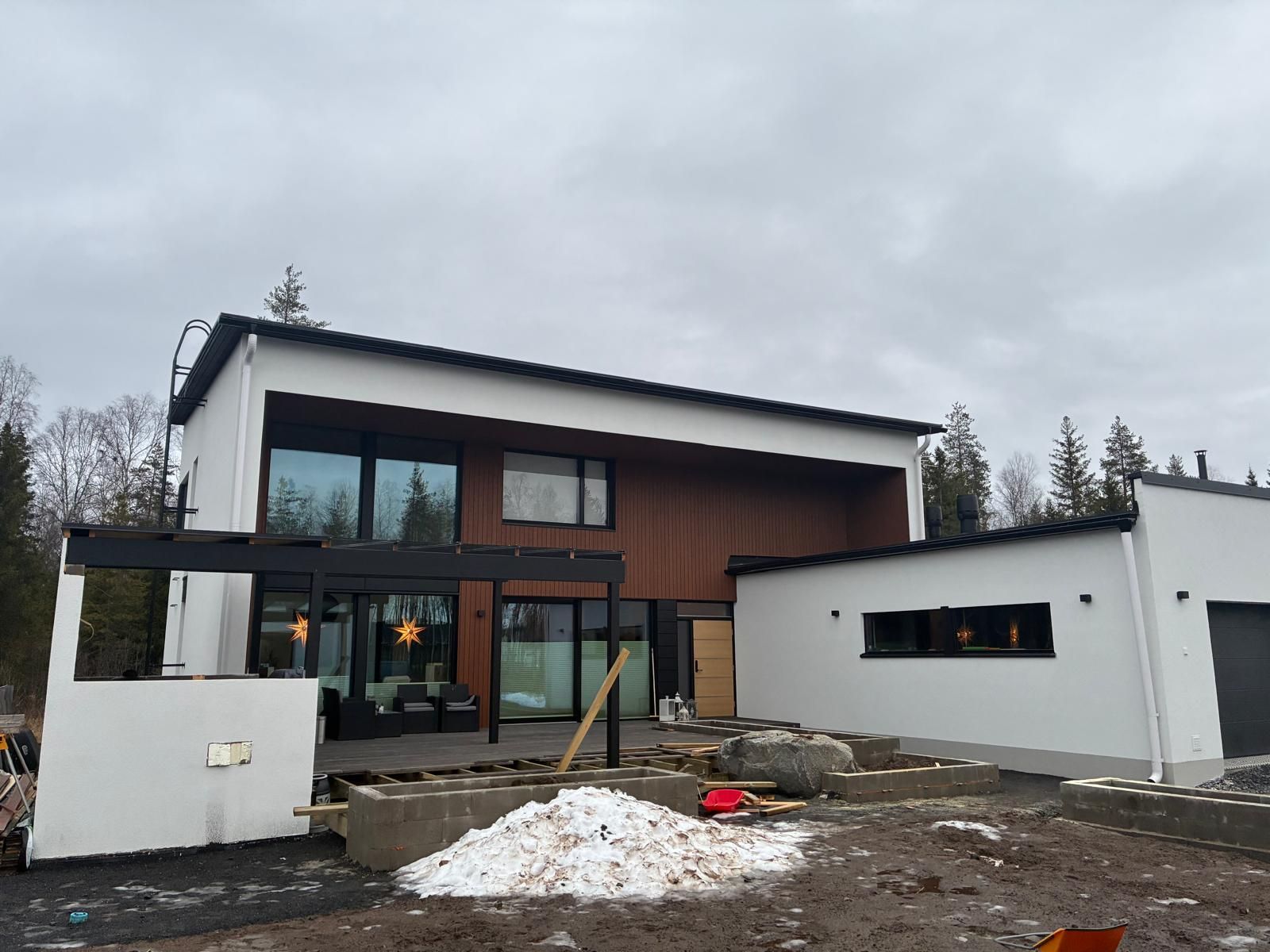 Moderni kaksikerroksinen koti, jossa on valkoiset ja puupaneloidut seinät, suuret ikkunat, musta pergola ja sorapäällysteinen piha.