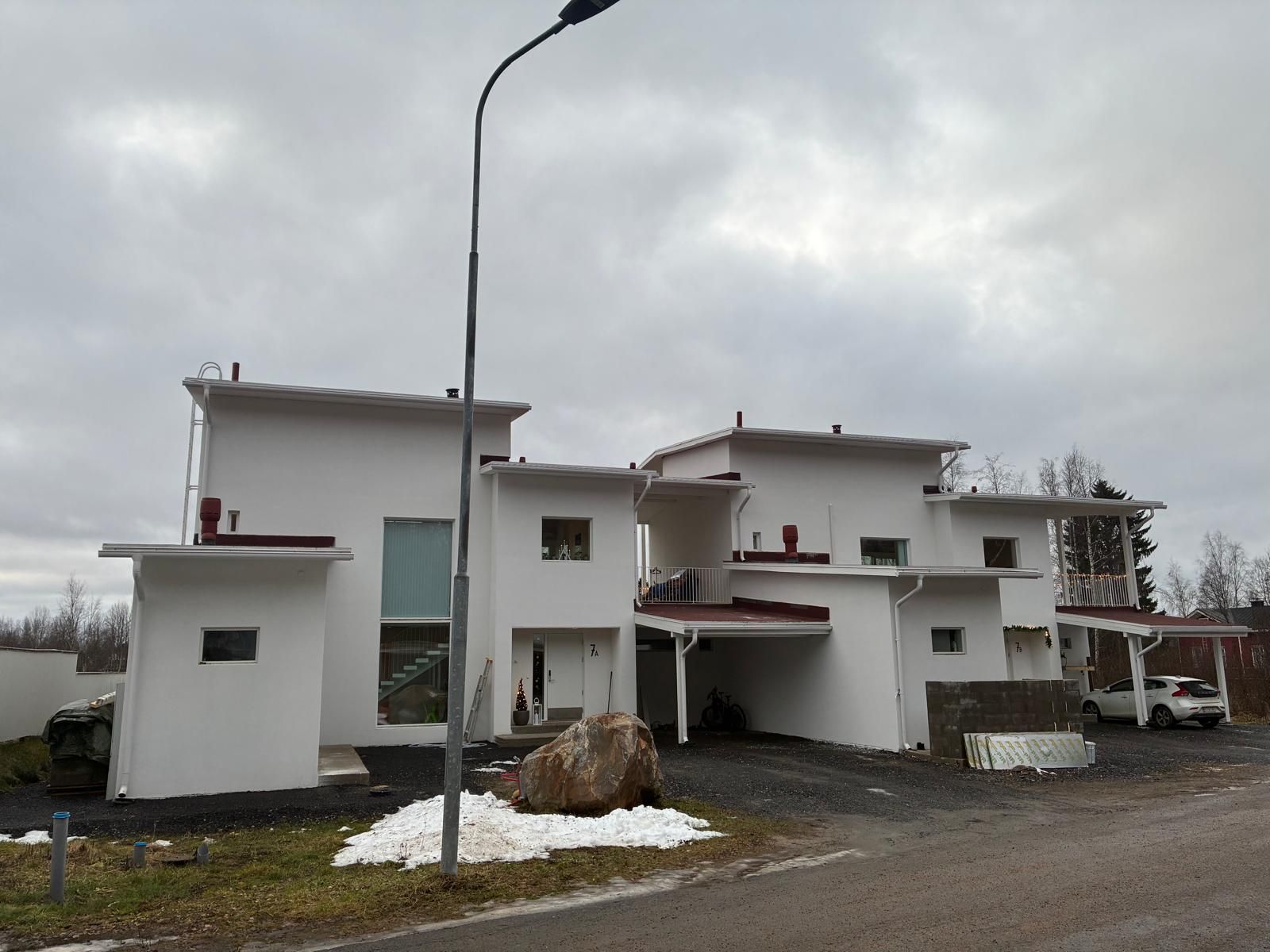Kaksi modernia valkoista taloa, joissa on tasainen katto ja autokatos, seisoo katuvalaisimen ja suuren lohkareen takana pilvisenä päivänä.