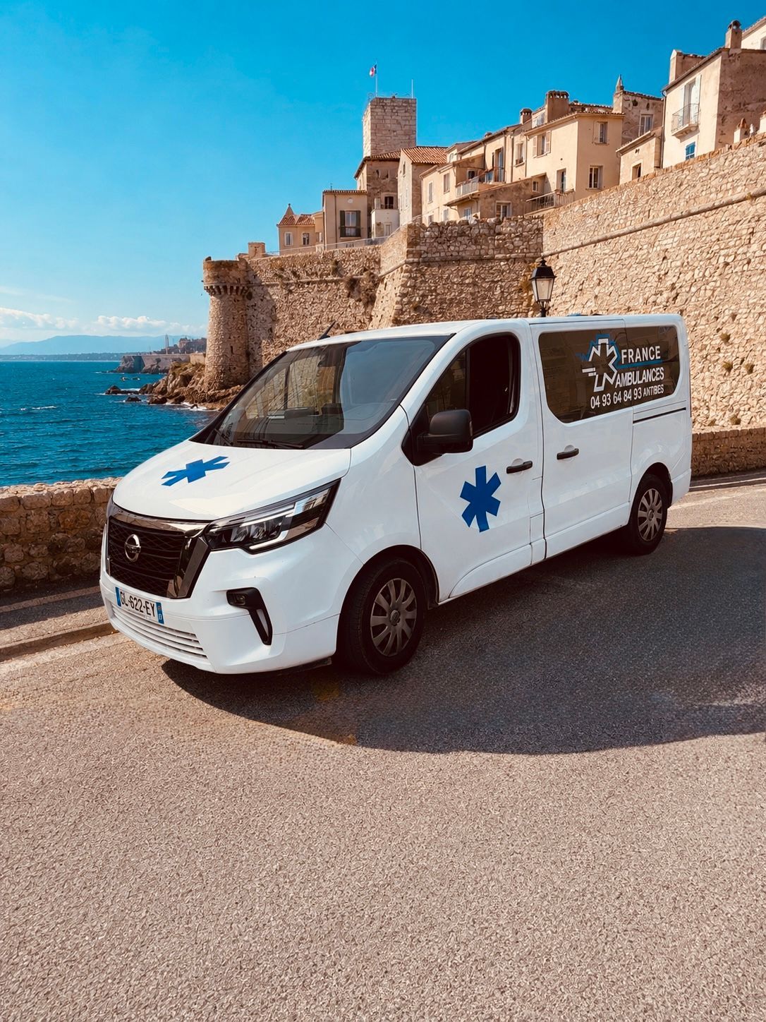 Une ambulance blanche est garée près d'un muret en pierre en bord de mer, avec des bâtiments en arrière-plan.