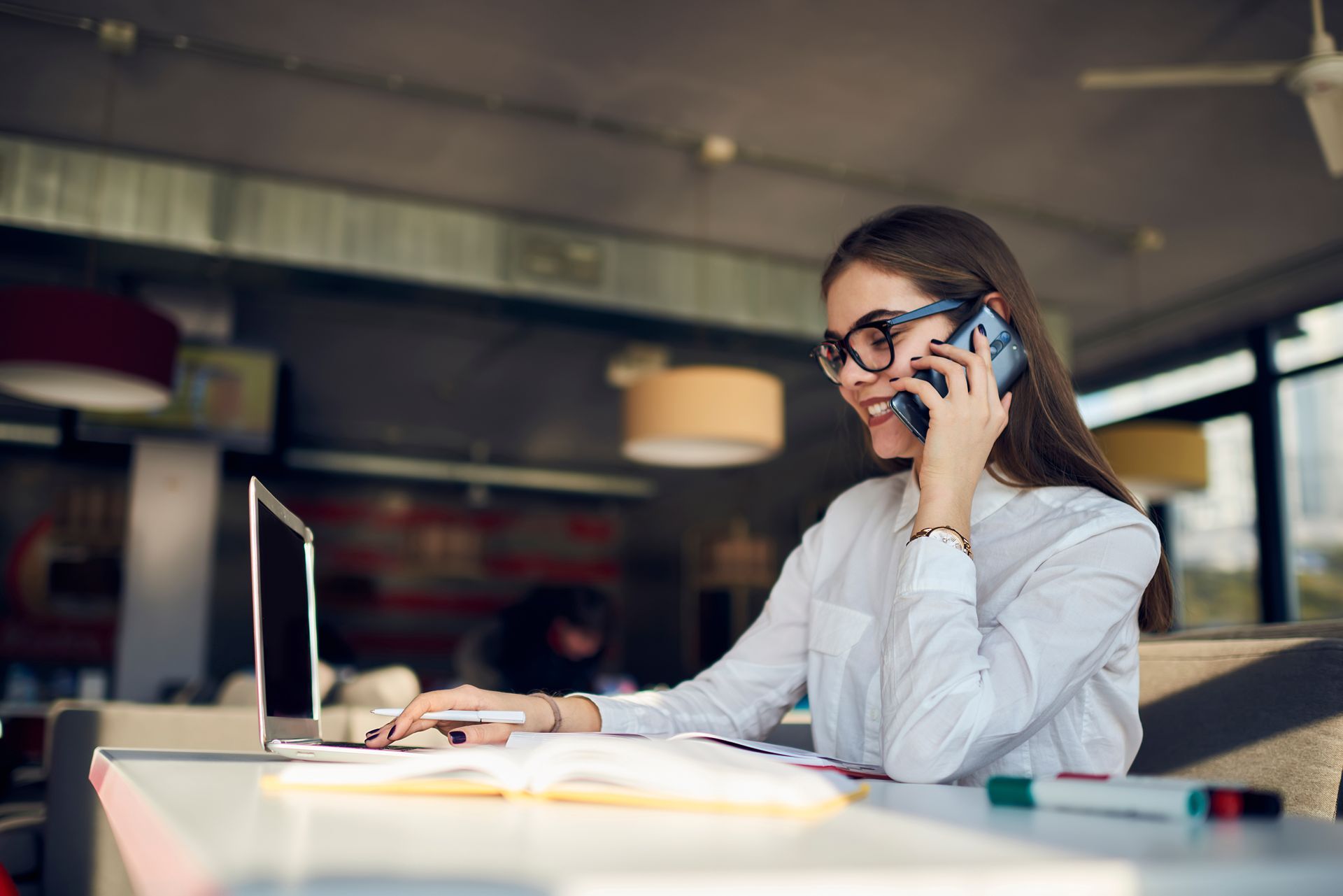 Une femme portant des lunettes travaille sur un ordinateur portable et parle au téléphone à la table d'un café.