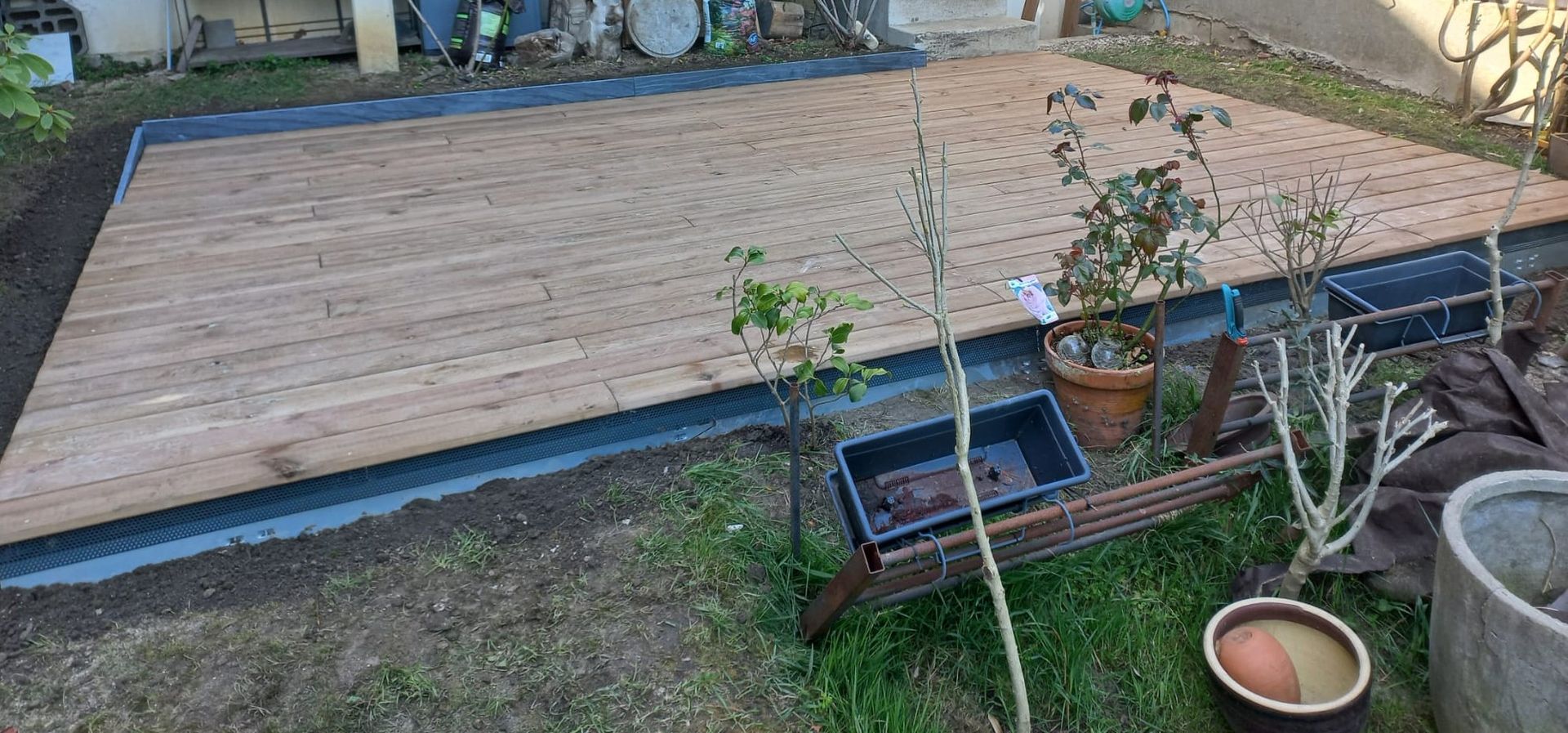 Terrasse en bois dans un jardin verdoyant, bordée de bordures noires. Plusieurs plantes en pot sont plantées à proximité.
