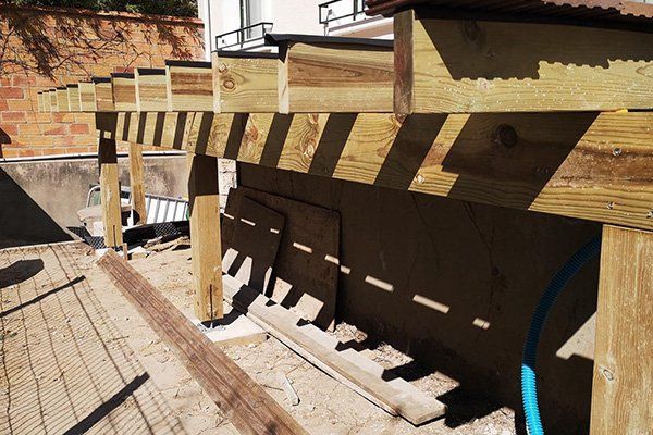 Terrasse en bois en construction, des poutres projetant des ombres sur un mur.