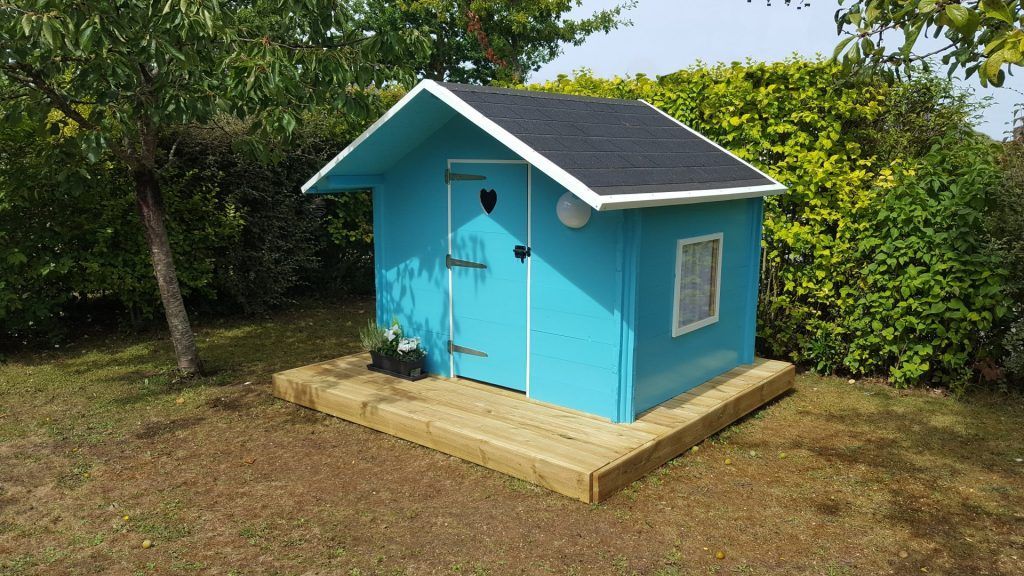 Maisonnette bleue sur plate-forme en bois dans une cour herbeuse.
