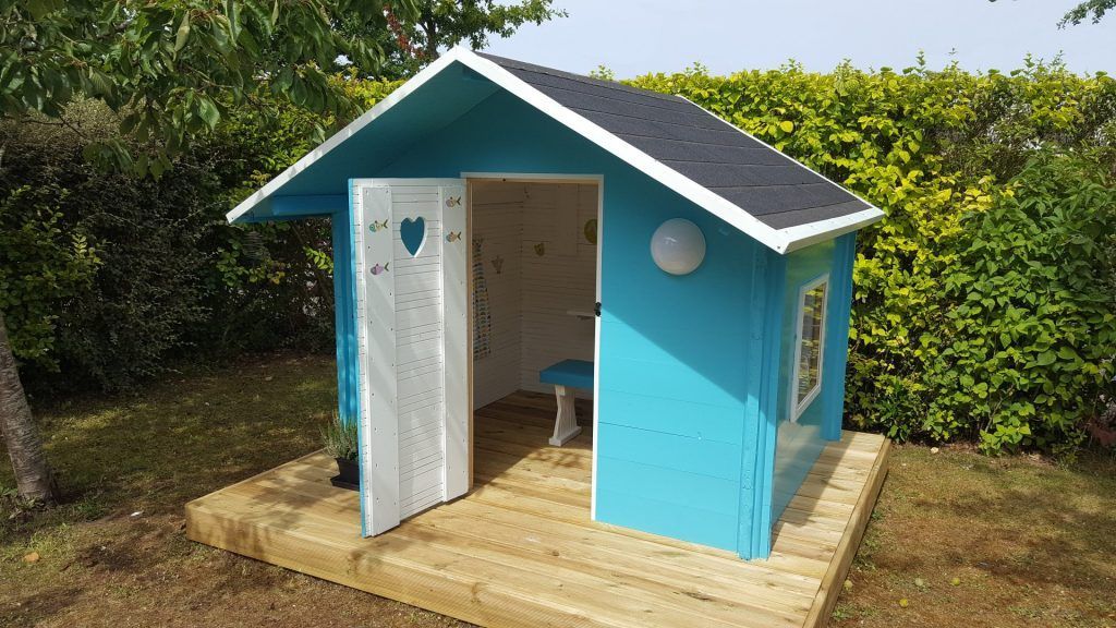 Maisonnette turquoise pour enfant avec porte ouverte sur une plate-forme en bois, entourée d'herbe et de buissons.