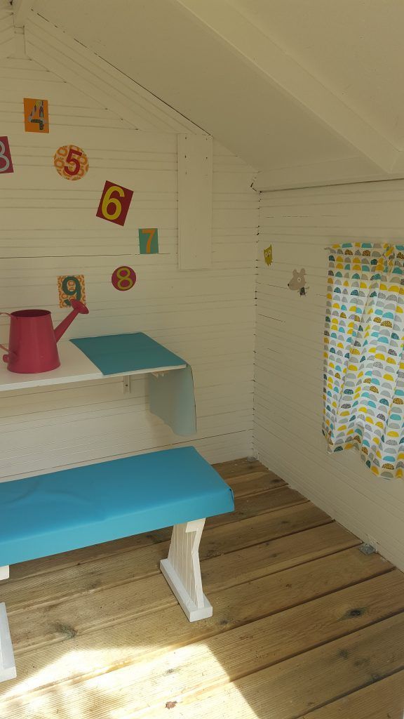 À l'intérieur d'une petite maison de jeu blanche avec une table de pique-nique bleue, un plancher en bois et des décorations de chiffres colorées.