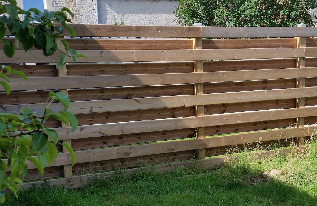 Clôture horizontale en bois dans le jardin. De l'herbe verte et du feuillage l'encadrent.