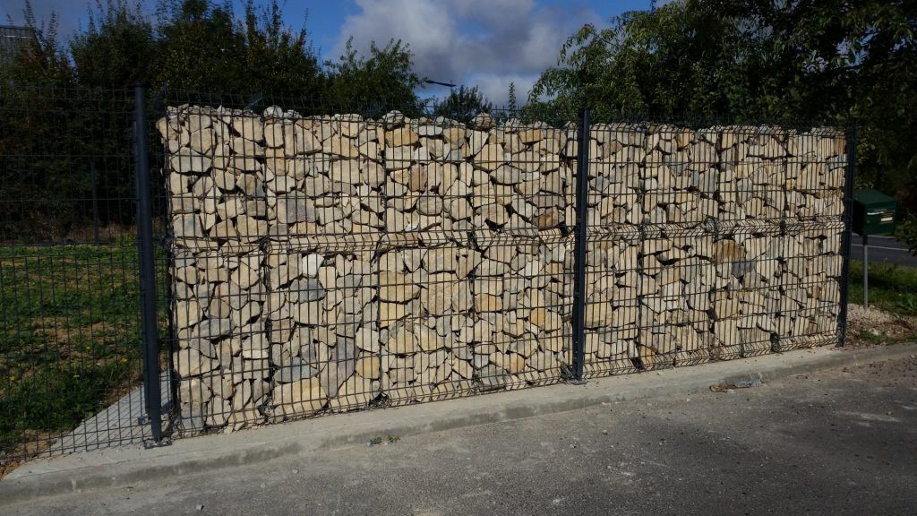 Mur de gabions rempli de pierres entre des poteaux métalliques, sur fond vert et ciel bleu.