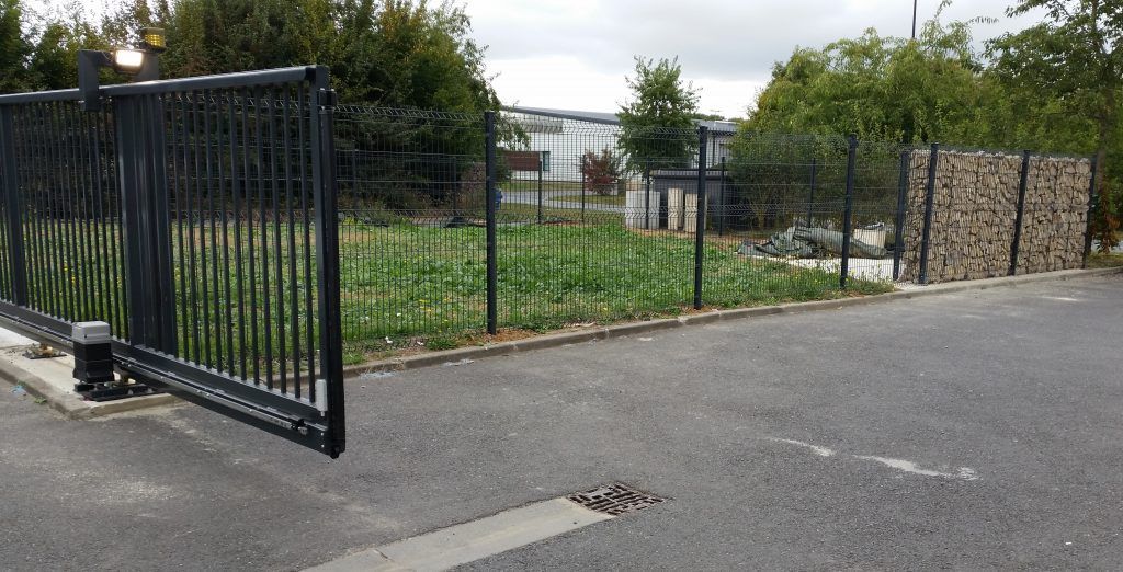 Portail en métal noir partiellement ouvert sur l'asphalte, menant à une zone herbeuse et des bâtiments, sous un ciel nuageux.