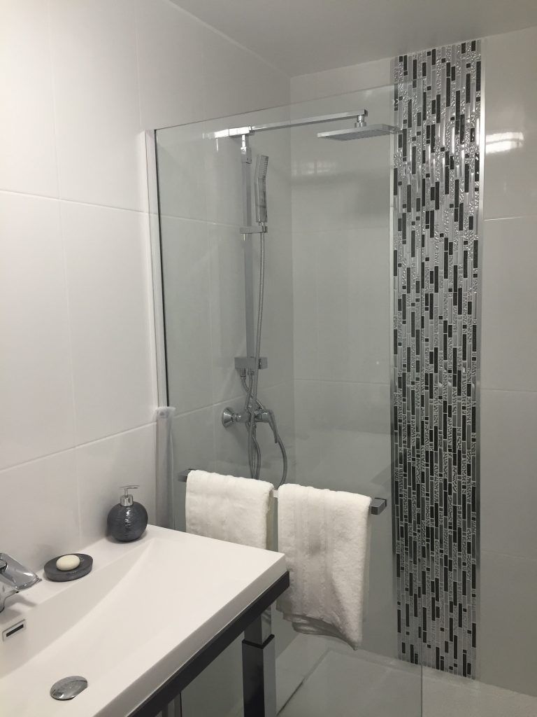 Salle de bain moderne avec carrelage blanc, douche en verre, robinetterie chromée et mosaïque. Lavabo et serviettes blancs.