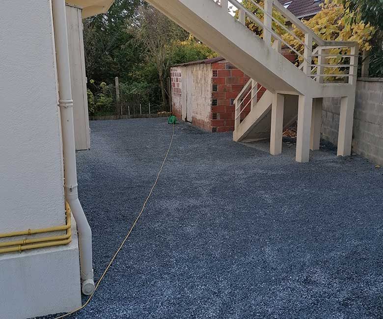 Cour de gravier sous les escaliers, un bâtiment à gauche, un hangar en briques et des arbres en arrière-plan.