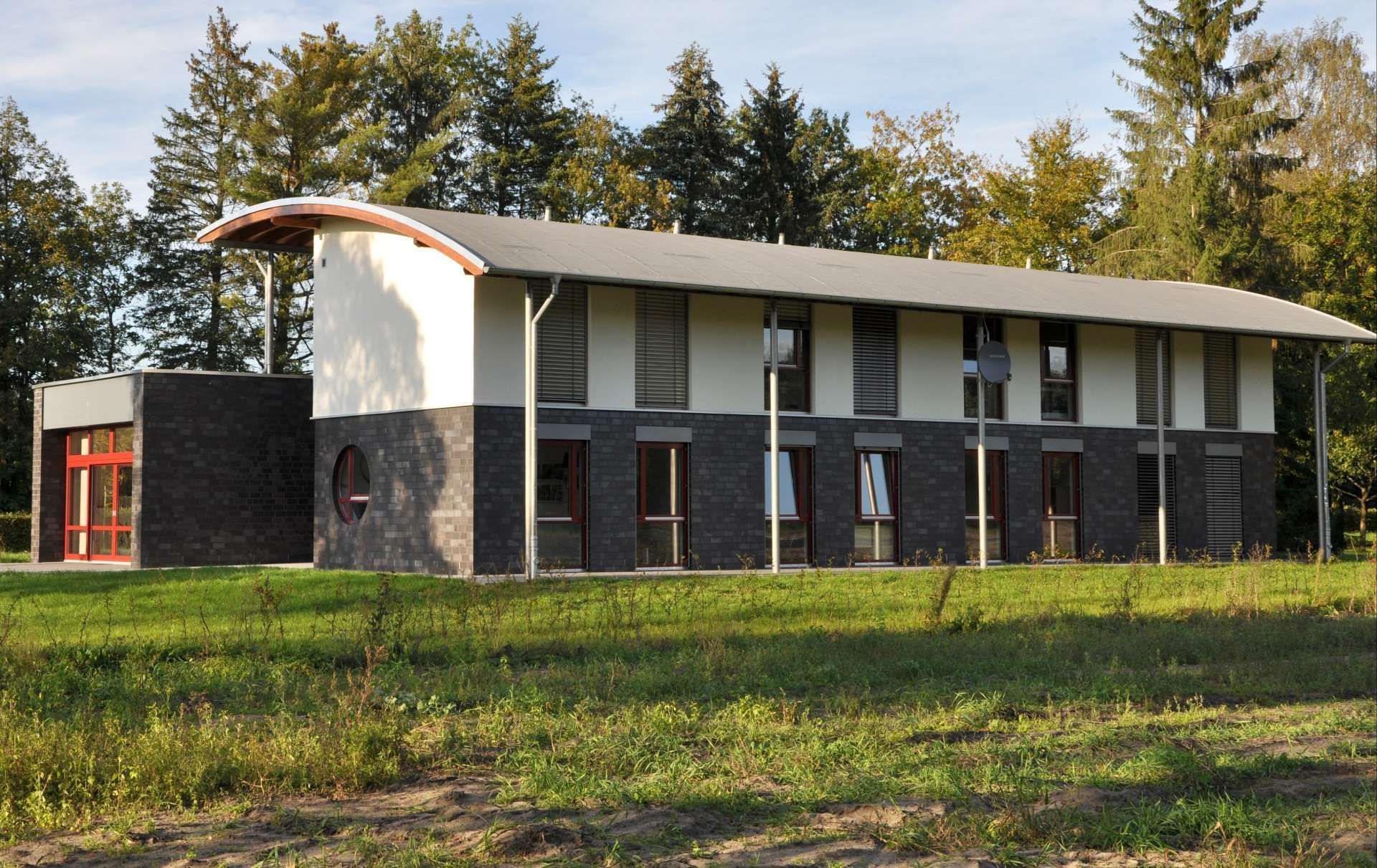 Modernes zweistöckiges Gebäude mit weißem, geschwungenem Dach und dunklen Backsteinmauern auf einem grasbewachsenen Feld